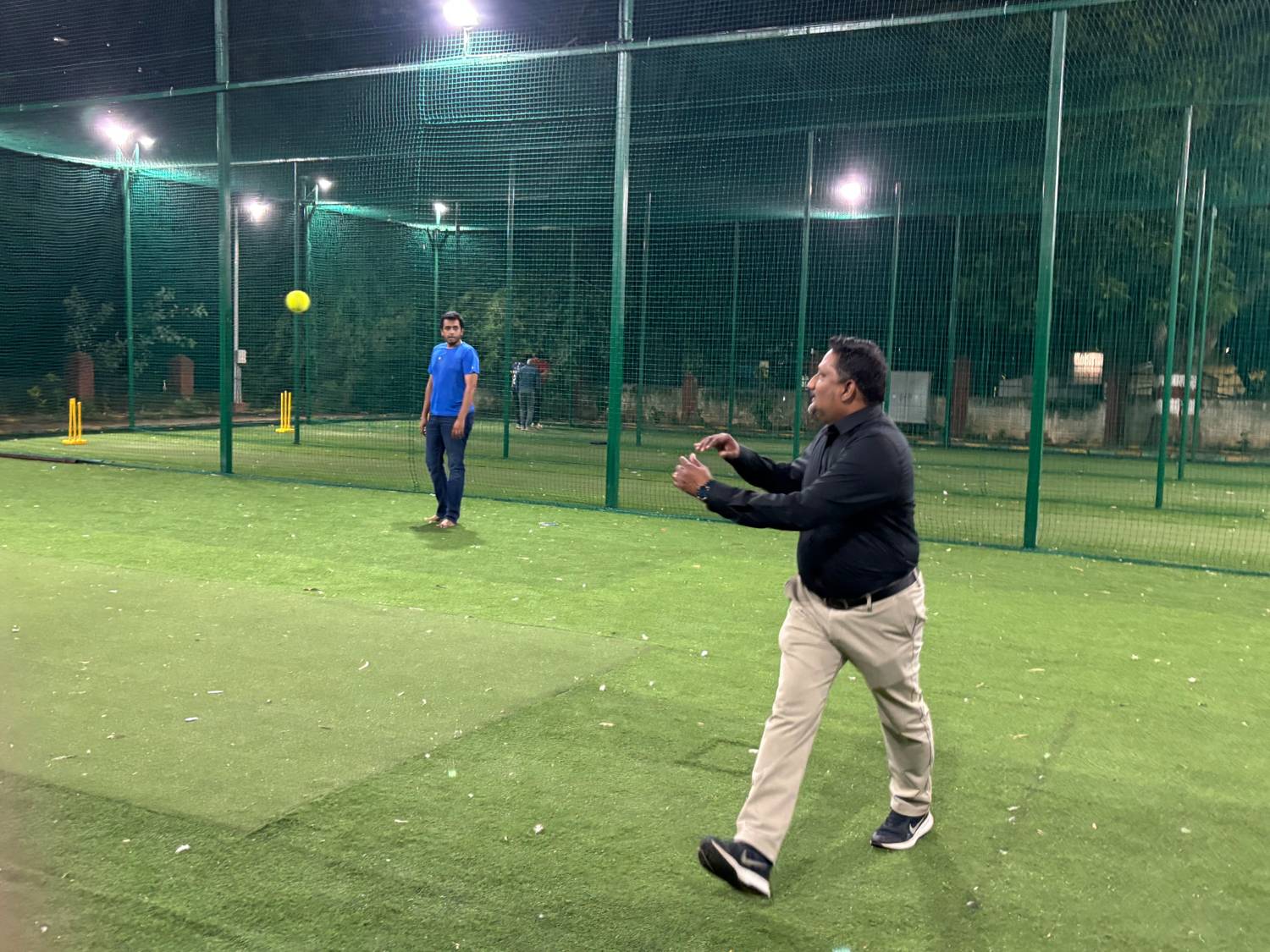 A tiramai team member bowling in the floodlit nets while a teammate waits at the far end